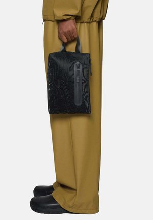 Persoon met een bruine broek en zwarte schoenen die een zwart gestructureerde zak met een verticale ritssluiting bij de handvatten vasthoudt.