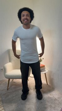 Uomo con acconciatura afro che indossa una maglietta bianca e pantaloni neri, sorride in una stanza con moquette con una sedia beige e un tavolino laterale.