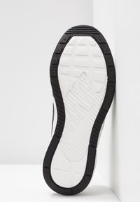 Mustat ja valkoiset tennarin pohjat, joissa on teksturoitu kumipinta, monisuuntaiset urat ja kohokuvioidut logo-yksityiskohdat.