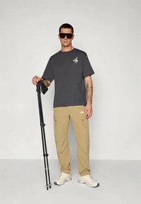 Άνδρας με γυαλιά ηλίου, σκούρο γκρι μπλουζάκι, μπεζ παντελόνι και λευκά αθλητικά παπούτσια, στέκεται κρατώντας μαύρα μπατόν πεζοπορίας μπροστά σε απλό φωτεινό φόντο.