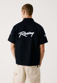 Melnas īsām piedurknēm kreklas ar apkakli, aizmugurē ar baltu tekstu "Racing". Gluda auduma standarta piegriezums. Minimalistisks dizains.