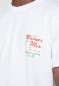 White cotton t-shirt featuring colorful text graphic, including "Mamma Mia" in red and green, with curved lettering and simple design.