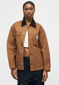 Sieviete valkā brūnu žaketi ar pogām un melnu apkakli, baltu īsā topu un tumši zilas bikses pret vienkāršu gaišu fonu.