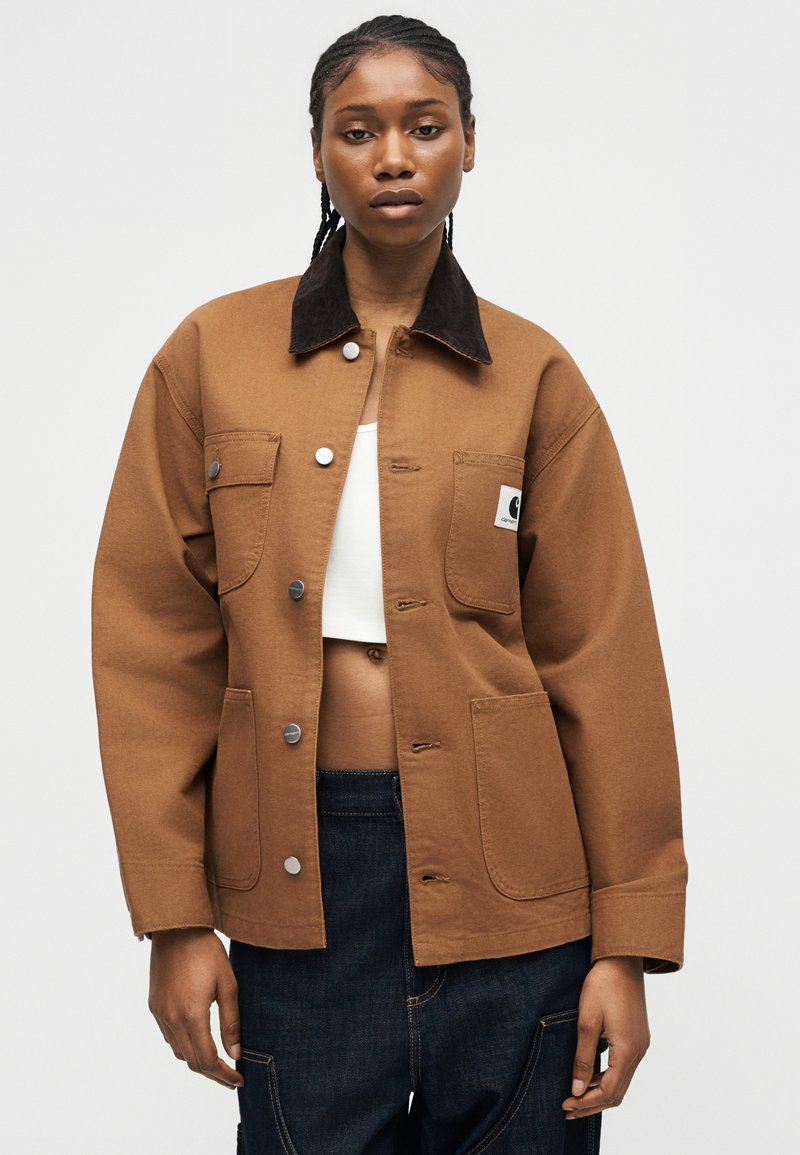 Donna che indossa una giacca marrone con bottoni e colletto nero, top bianco corto e pantaloni blu scuro su uno sfondo chiaro e uniforme.