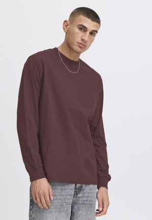 Giovane uomo con capelli corti che indossa una camicia bordeaux a maniche lunghe, una collana con catena d'argento e jeans grigio chiaro su uno sfondo semplice.