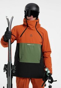 Orangen- und schwarze, wasserdichte Skijacke mit grünen Akzenten, Reißverschluss-Tasche und verstellbarem Kapuze. Glänzender schwarzer Helm und Skistöcke.