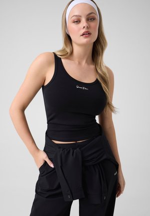 Jeune femme aux longs cheveux blonds portant un bandeau blanc, un haut noir sans manches et un pantalon noir avec une veste nouée autour de la taille.