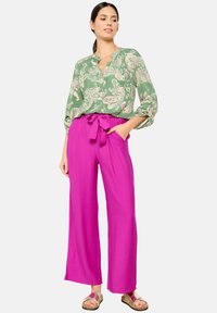 Blusa verde con motivo paisley e scollatura a V, abbinata a pantaloni larghi rosa acceso con cinturino annodato. Sandali color sabbia completano l'outfit.