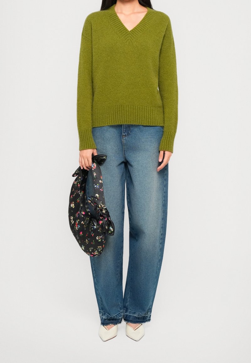 Personne portant un pull à col en V vert olive, un jean droit bleu, des chaussures blanches à bout pointu, tenant un sac noir à motif floral.
