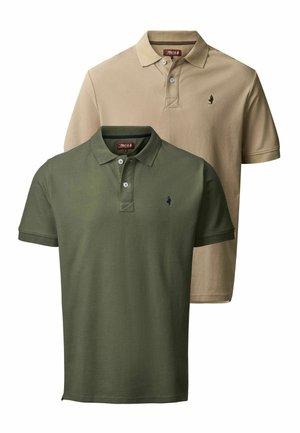 Homme avec des tatouages portant un polo vert olive, un jean noir et des bottes en cuir marron, debout devant un fond blanc uni.