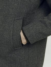 Manteau en chevron gris avec une poche latérale et une doublure beige qui dépasse du poignet, mettant en valeur un tissu en mélange de laine texturé.