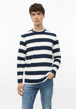 Jonge man die een marineblauw-wit horizontaal gestreept shirt met lange mouwen en blauwe jeans draagt, staat met één hand in de zak.