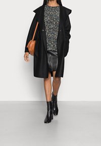 Schwarzer Wollmantel, gemustertes dunkles Oberteil, Lederrock und schwarze Stiefel mit Absatz. Braune Handtasche in runder Form. Flacher, grauer Hintergrund.