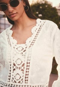 White embroidered blouse featuring intricate lace details and cut-outs. The neckline has scalloped edges, and the fabric appears lightweight.