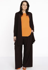 Sort lang jakke over en orange top, parret med sorte brede bukser og orange sko. Outfitet har et lagdelt design med en flydende tekstur.