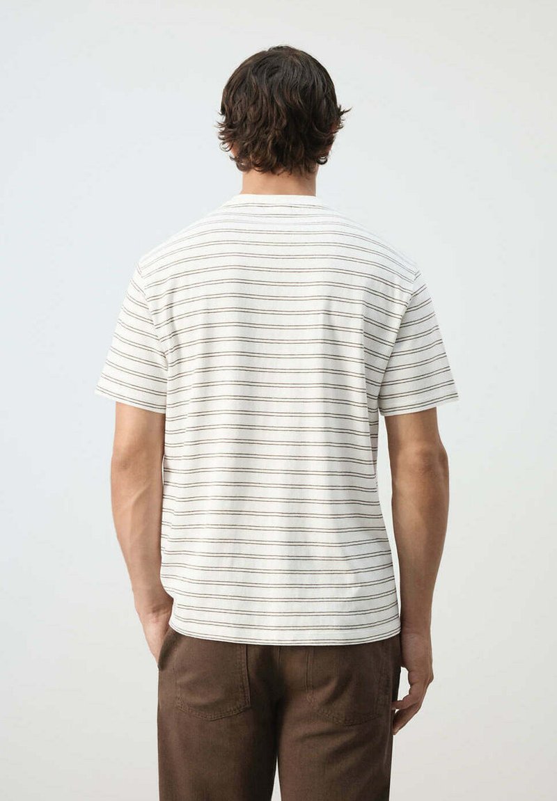 Hombre con cabello corto y oscuro que lleva una camisa blanca de manga corta con finas rayas horizontales y pantalones marrones, visto desde atrás.