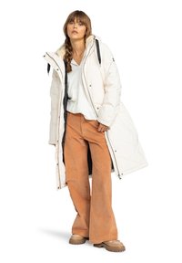 Manteau puffer long blanc avec une capuche en fourrure, fermeture éclair à l'avant et accents noirs ; associé à un pantalon large en velours côtelé orange et des bottes beiges.