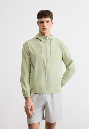 Νέος άνδρας με ανοιχτό πράσινο φούτερ με φερμουάρ και κουκούλα και ανοιχτό γκρι σορτς, στέκεται μπροστά σε λευκό φόντο.