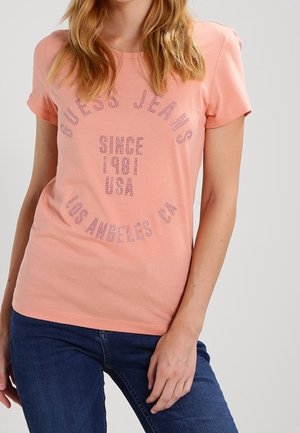 Femme portant un t-shirt à manches courtes couleur pêche avec l'inscription « Guess Jeans Since 1981 USA Los Angeles CA » en strass roses, associé à un jean bleu.