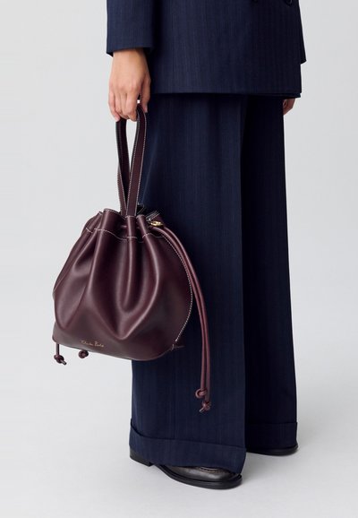 Borsa a secchiello in pelle marrone con chiusura a coulisse, caratterizzata da cuciture a contrasto e un logo dorato. Indossata al braccio con pantaloni larghi in navy.