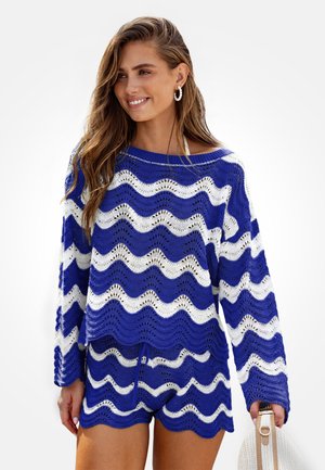 Donna con maglione a righe ondulate blu e bianco abbinato a pantaloncini coordinati, che tiene una borsa tote intrecciata bianca.