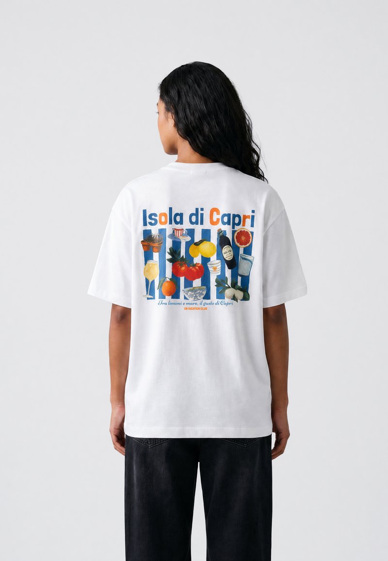 Άτομο με μακριά σκούρα μαλλιά φοράει λευκό t-shirt με πολύχρωμο τύπωμα Isola di Capri που παρουσιάζει εικονογραφήσεις φαγητού και ποτών στην πλάτη.