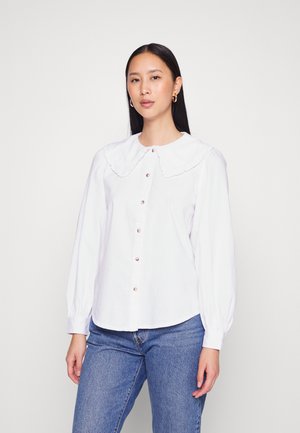 Blusa branca de mangas longas com um grande colarinho volantado, fechamento frontal com botões e textura suave, acompanhada de jeans de ganga azul claro.