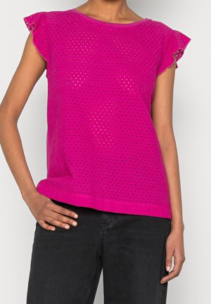Woman wearing a bright pink, perforated short-sleeve top with ruffled sleeves, paired with black pants, hand resting on hip.
