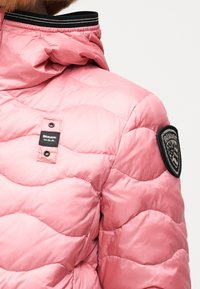 Ροζ μπουφάν με πλούσιο καπιτονέ, διπλωμένο γιακά, μαύρη και λευκή ριγέ λεπτομ έρεια, μαύρη ετικέτα με την επιγραφή "Blauer U.S.A." και εμβληματικό μπαλώμα στο μανίκι.