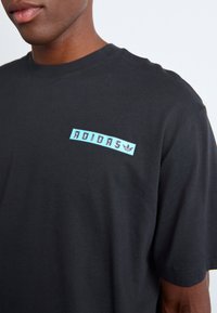 Sort bomulds t-shirt med et lysblåt "ADIDAS" logo og et trefoil accent i det øverste venstre brystområde. Korte ærmer og rund hals.