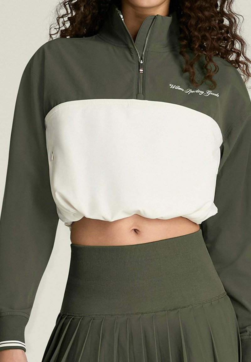 Frau trägt ein olivgrünes und weißes, kurz geschnittenes Pullover mit Reißverschluss und einen hoch taillierten, olivgrünen, plissierten Rock.