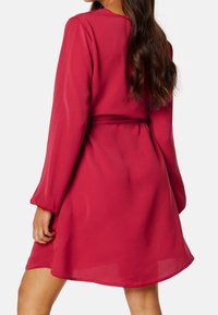 Robe rouge à manches longues en tissu léger, taille cintrée avec un nœud, ourlet fluide et texture légèrement transparente.