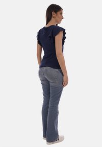 Donna vista di profilo che indossa una maglia blu scuro con maniche arricciate, jeans blu, sneakers bianche e orecchini a cerchio dorati su sfondo bianco.