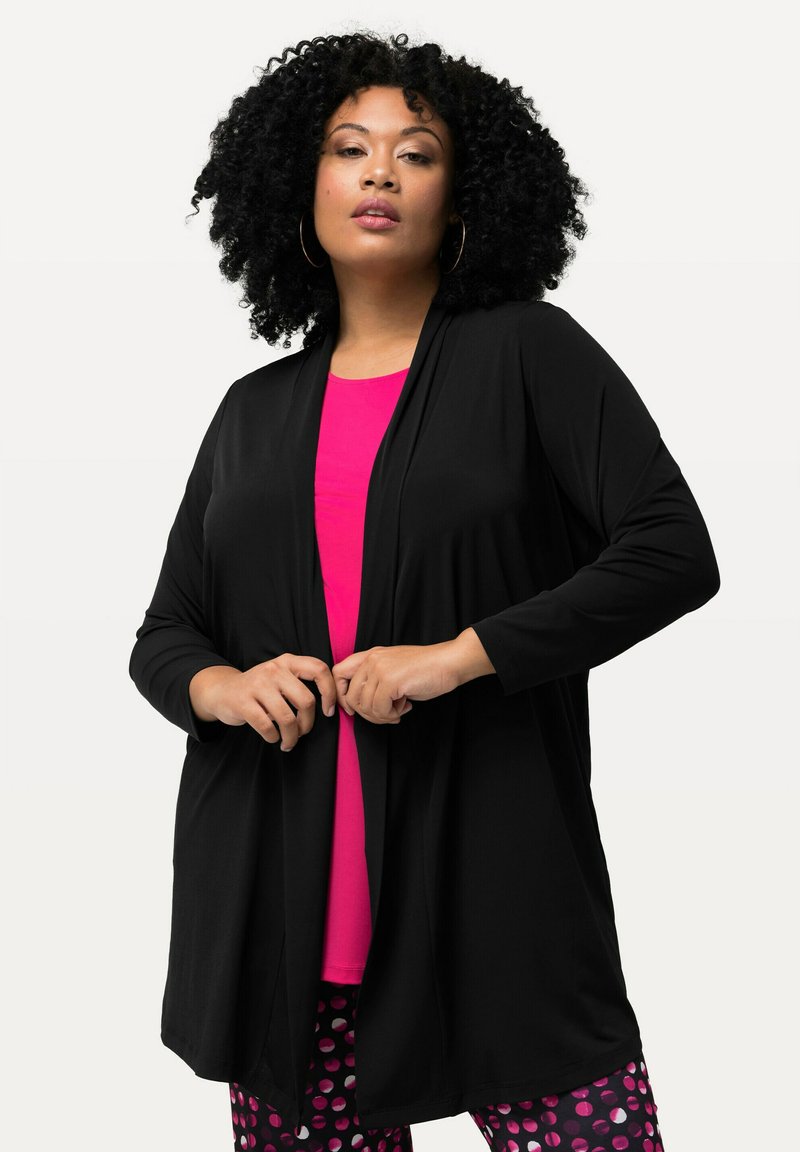 Schwarze offene Strickjacke aus weichem Material, kombiniert mit einem pinken Oberteil und gemusterten Hosen mit Kreisen in verschiedenen Farben.