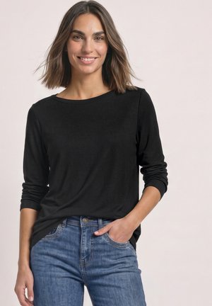 Femme souriante portant un t-shirt noir à manches longues et un jean bleu, une main dans la poche, sur un fond clair uni.