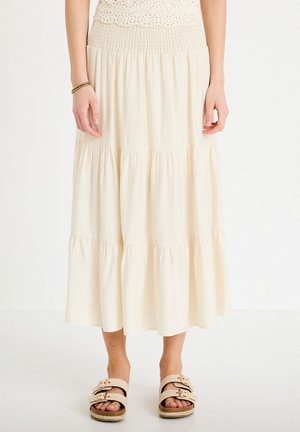 Femme portant une jupe midi crème à volants avec taille smockée, un haut en crochet, un bracelet en or et des sandales beiges à double boucle, debout sur un sol blanc.