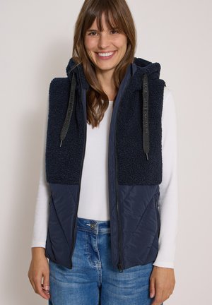 Donna sorridente che indossa un gilet senza maniche blu navy con pile e tessuto trapuntato, camicia bianca a maniche lunghe e jeans blu.