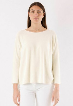 Donna con capelli lunghi e scuri, che indossa un maglione crema largo e pantaloni bianchi, in piedi davanti a uno sfondo chiaro e uniforme.