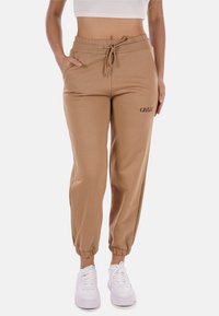 I pantaloni da tuta beige con cintura elastica presentano un cordoncino, polsini alle caviglie e la parola "GILLE" ricamata in nero sulla coscia sinistra.