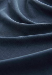 Námořnická modrá látka s hladkou texturou, vykazující jemné záhyby a odrazy světla, což naznačuje měkký a ohebný materiál.