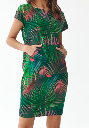Femme portant une robe verte avec un imprimé feuilles tropicales et oiseaux rose et vert, debout les mains dans les poches de la robe sur fond blanc.