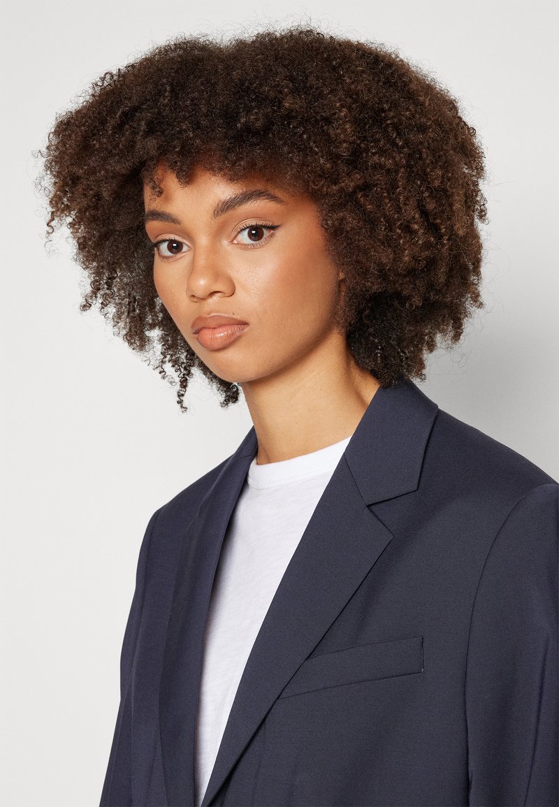 Blazer marine avec un col élégant, une poche unique et une texture lisse, porté sur un T-shirt blanc. Le modèle a les cheveux courts et bouclés.
