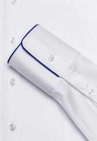 Chemise blanche avec un col à accents bleus, dotée de boutons en nacre et d'une texture lisse. Le poignet est replié pour révéler une bordure bleue.