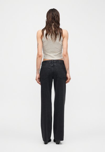 Femme aux cheveux longs et ondulés, portant un haut sans manches beige à rayures et un jean noir taille haute à jambes droites, debout, vue de dos.