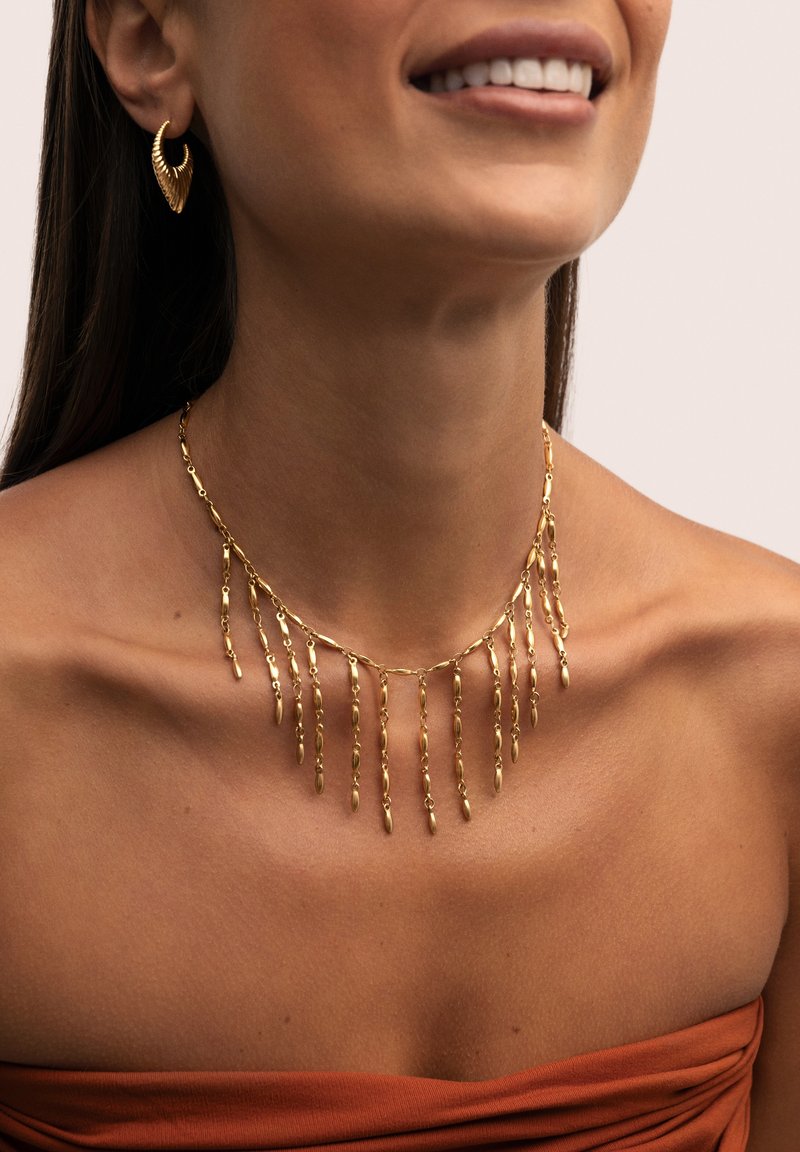 Collana dichiarativa in oro con dettagli pendenti a catena. Grandi orecchini ad anello texturizzati completano il design. Texture della pelle liscia visibile.