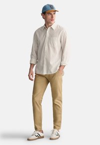 Lys beige stribet skjorte med knapper, tan slim-fit bukser og hvide sneakers med sorte striber. Lys blå cap med en tan skygge.