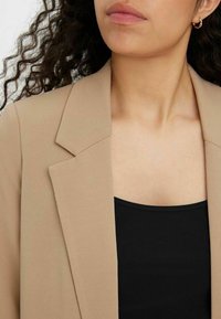 Blazer beige con un tessuto testurizzato, rever affilati e una vestibilità rilassata, indossato sopra un top nero aderente. Orecchini a cerchio dorati visibili.