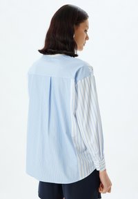 Camisa de color azul claro con un yugo sólido y mangas a rayas, con un corte relajado, puños abotonados y un pliegue en la parte posterior para un detalle adicional.