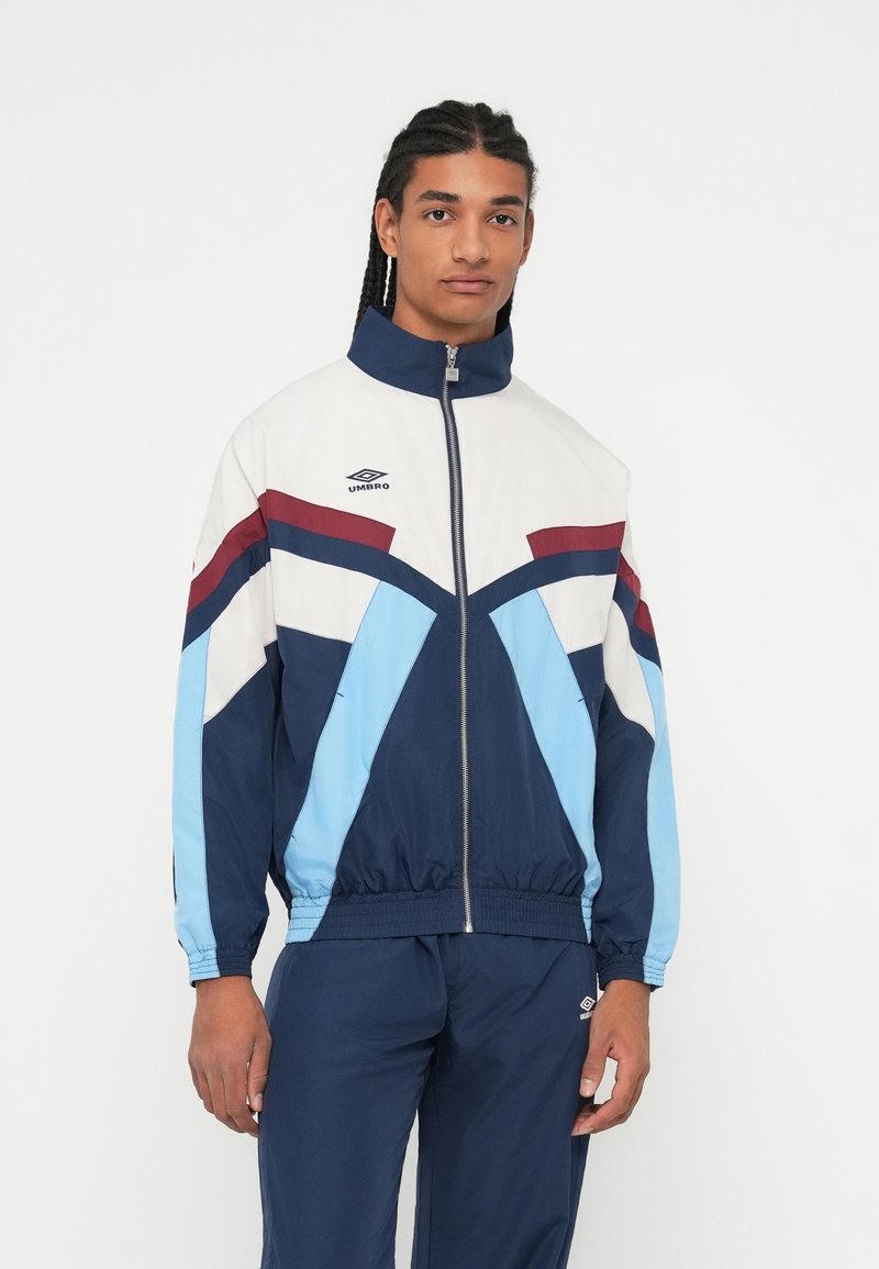 Young man wearing a blue, white, and maroon Umbro track jacket with matching navy track pants, standing against a plain white background.