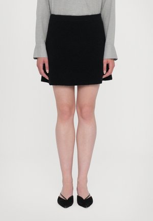 Femme portant une mini-jupe noire, un haut gris à manches longues avec des poignets évasés, et des talons noirs pointus sur un fond blanc uni.